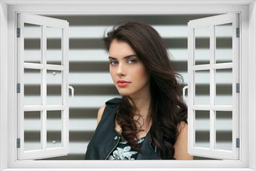 Closeup portrait of young beautiful brunette woman in black leather jacket posing outdoors against urban style background of metal strips