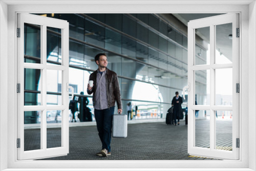 Portrait of a young man walking near the airport with suitcase and coffee