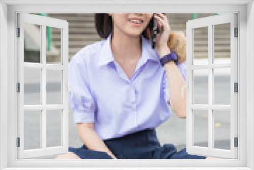 Cute Asian Thai high schoolgirl student in school uniform sitting and talking with her smart phone showing fun smiling happy facial expression