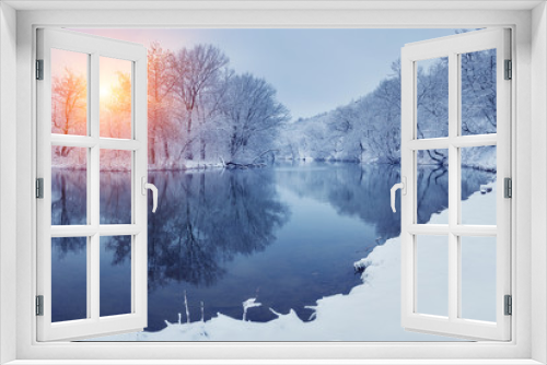 Fototapeta Naklejka Na Ścianę Okno 3D - Winter forest on the river at sunset. Panoramic landscape with snowy trees, sun, beautiful frozen river with reflection in water. Seasonal. Winter trees, lake and blue sky. Frosty snowy river. Weather