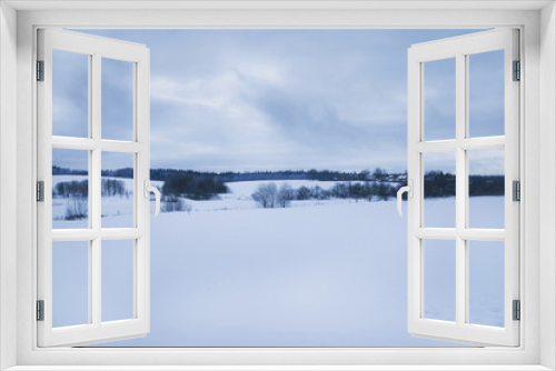 Fototapeta Naklejka Na Ścianę Okno 3D - Snow-covered fields and hills, forest and village in the background, a gray-blue cloudy sky