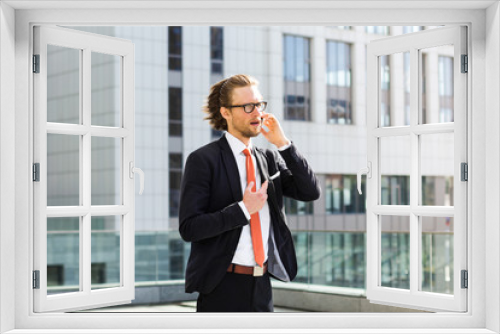 A businessman with a beard and glasses talking on the phone on the background of the building.