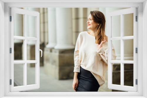 Slim elegant woman with red lips in a beige coat and black skirt brunette with long legs near the columns.