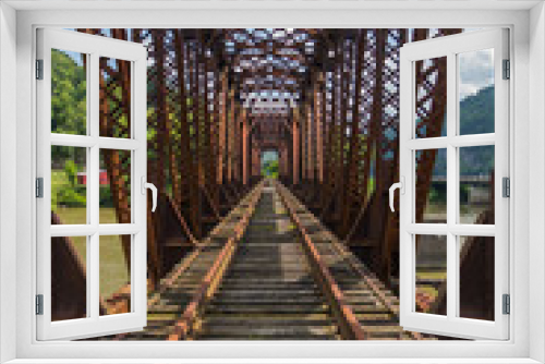 Fototapeta Naklejka Na Ścianę Okno 3D - A down the track perspective across an old rusted railroad bridge spanning a river with green mountains in the distance with a blue cloudy sky.