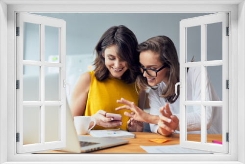 Two joyful coworkers browsing phone laughing together while taking break from work in the office