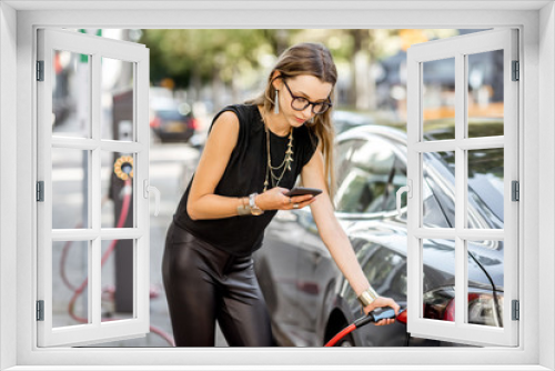 Young woman charging electric car standing with smart phone outdoors on the street in Rotterdam city