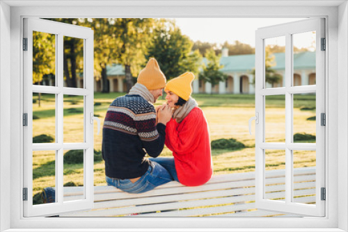Fototapeta Naklejka Na Ścianę Okno 3D - Romantic couple sit on bench, enjoy sunny day, keep hands together, look with great love at each other, have good relationships. Female has date with boyfriend in park, admire beautiful nature