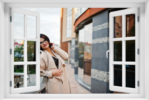 Black hair sexy woman in glasses and coat posed against building with modern windows.