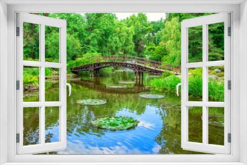 Fototapeta Naklejka Na Ścianę Okno 3D - View of beautiful garden with wooden walking bridge, green trees, bushes and blue sky, reflecting in a pond water. Summer natural landscape