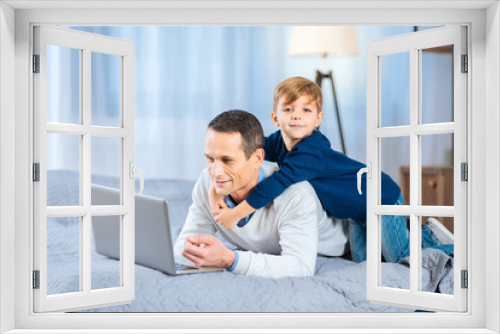 Gentle hug. Adorable little boy back-hugging his beloved father lying on the bed and working on the laptop