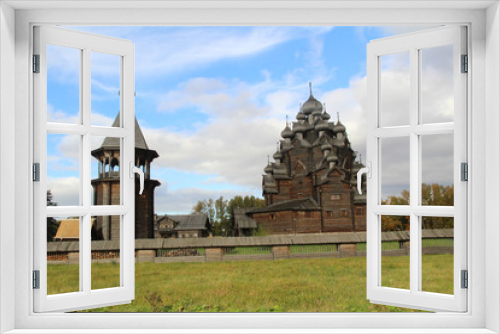 Fototapeta Naklejka Na Ścianę Okno 3D - The bell tower from logs and traditional Russian church from logs, surrounded with a fencing from logs against the background of the bright autumn sky.