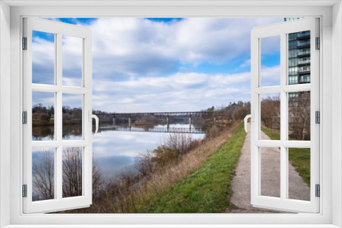 Fototapeta Naklejka Na Ścianę Okno 3D - Narrow Path alongside a River and Blue Sky with Clouds on a Late Autumn Day. An Old  Railway Bridge Crossing the River in Visible in Background. Reflection in Water.