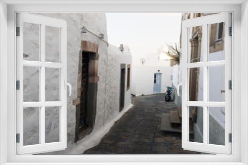 Fototapeta Naklejka Na Ścianę Okno 3D - A view of a narrow street with arch and wooden windows and doors with white wall stone architecture of the island Patmos, Greece
