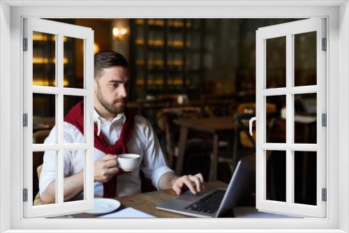 Serious businessman with drink sitting by table in front of laptop and searching for necessary data