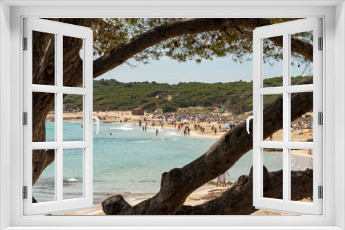 Fototapeta Naklejka Na Ścianę Okno 3D - View through the branches of a tree to the beach and the Mediterranean Sea with visitors from Cala Agulla with a wooden hut on the Spanish holiday island Mallorca