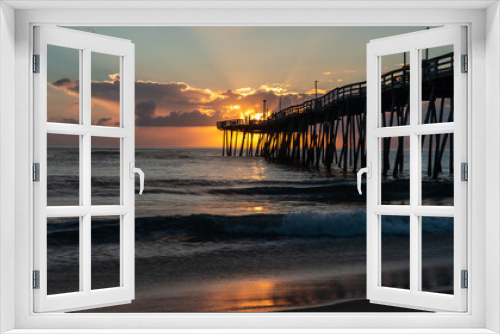 Fototapeta Naklejka Na Ścianę Okno 3D - The golden light of a bright sunrise beams through the clouds to light up a fishing pier that extends out into the ocean. Small waves break on the sandy shore. Sunrise colors reflected on the surface.