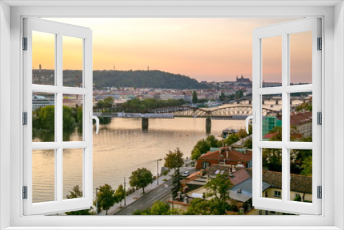 Fototapeta Naklejka Na Ścianę Okno 3D - View from Vysehrad of Prague castle, sunset sky with pink and yellow colors, railway bridge, stone bridge, river Vltava, boats, riverbank, streets, cars, houses, red roofs, green trees, hill of Petrin
