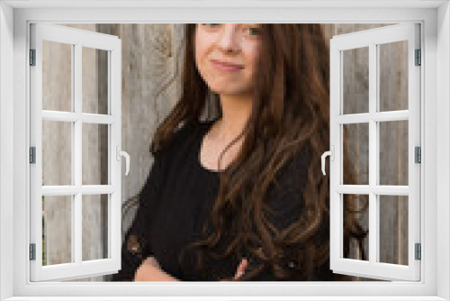 Brunette woman posing in front of a rustic old wooden fence wearing a black dress standing with arms crossed. Portrait of a young white woman standing in front of a wood fence outside in the summer.