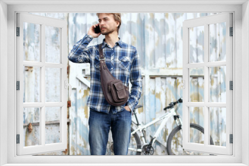 Hipster bearded man with modern leather bag talks on smartphone. Grunge city wall with bicycle on background. Modern city lifestyle concept