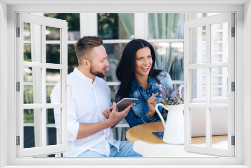 The concept of gadgets, technology, freelancing, people. A young European man sits at a table in a cafe with his girlfriend and talks about something, laughs. Friends rest in the restaurant outside