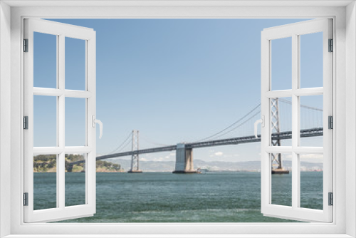 Fototapeta Naklejka Na Ścianę Okno 3D - A beautiful view of the expensive and famous San Francisco Oakland Bay Bridge with suspension cables and towers protruding from the San Francisco Bay with water below and clear blue sunny sky above.