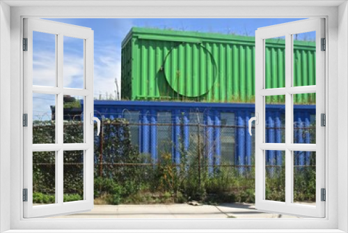 Fototapeta Naklejka Na Ścianę Okno 3D - Green container stacked on a blue container behind a chain link fence, with vegetation growing on and around them.