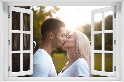 Happy young cute loving couple standing near tree in park kissing with beautiful sundown light.