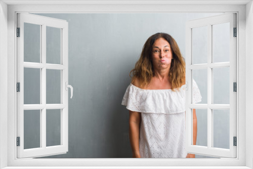 Middle age hispanic woman standing over grey grunge wall puffing cheeks with funny face. Mouth inflated with air, crazy expression.