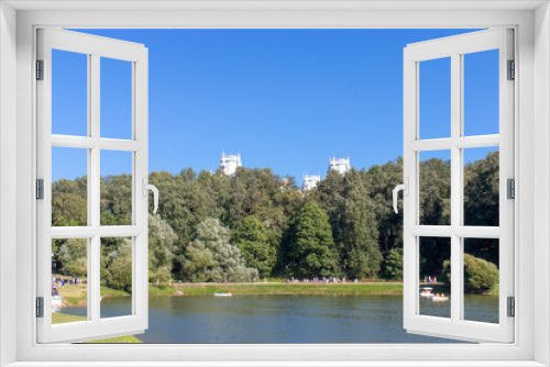 Fototapeta Naklejka Na Ścianę Okno 3D - Pond with green trees on the shore. The towers of the building in the blue sky, peeking from the trees. People boating on the lake and walk along the shore.