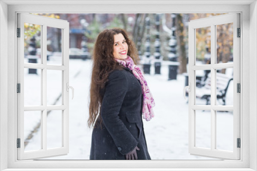 City, winter and people concept - Beautiful happy young woman in black coat and pink scarf stands sideways in snowy park