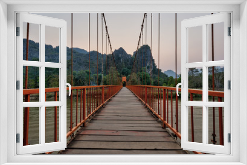 Fototapeta Naklejka Na Ścianę Okno 3D - Orange suspension footbridge over the Nam Song River and scenic limestone karst mountains near the Tham Chang (or Jang or Jung) Cave in Vang Vieng, Laos, at sunset.