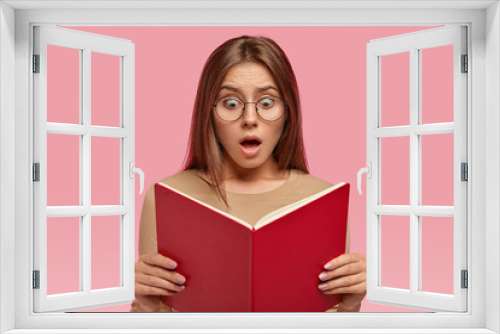 Headshot of surprised woman with terrified expression, stares at book, reads shocking information while prepares for examination, keeps jaw dropped, isolated over pink background. Reaction concept