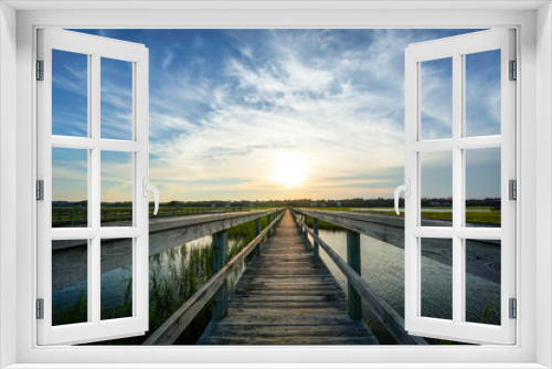 Fototapeta Naklejka Na Ścianę Okno 3D - coastal waters with a very long wooden boardwalk pier in the center during a colorful summer sunset under an expressive sky with reflections in the water and marsh grass in the foreground