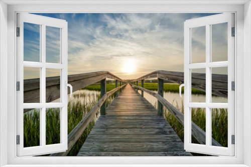 Fototapeta Naklejka Na Ścianę Okno 3D - coastal waters with a very long wooden boardwalk pier in the center during a colorful summer sunset under an expressive sky with reflections in the water and marsh grass in the foreground