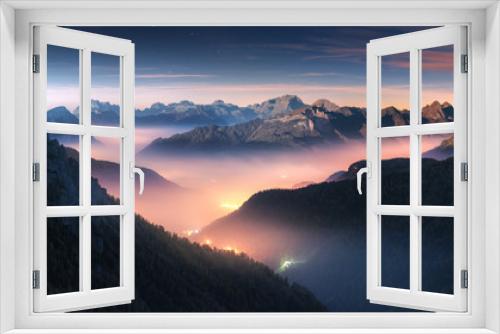 Fototapeta Naklejka Na Ścianę Okno 3D - Mountains in fog at beautiful night in autumn in Dolomites, Italy. Landscape with alpine mountain valley, low clouds, forest, colorful sky with stars, city illumination at dusk. Aerial. Passo Giau