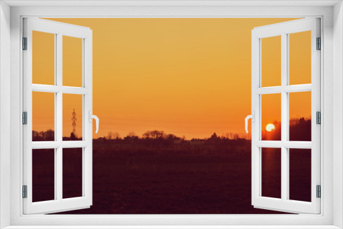 Fototapeta Naklejka Na Ścianę Okno 3D - Sunset landscape, orange sky, sun setting down, dark horizon with silhouettes of electricity pylon, wires, houses and trees, rural countryside