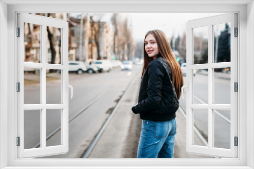 Portrait from back of elegant fashion girl long hair walking on on city background. She has black leather jacket and jeans. She is smiling to camera.