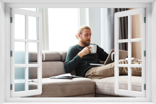 Concentrated young bearded man sitting in home using laptop computer.