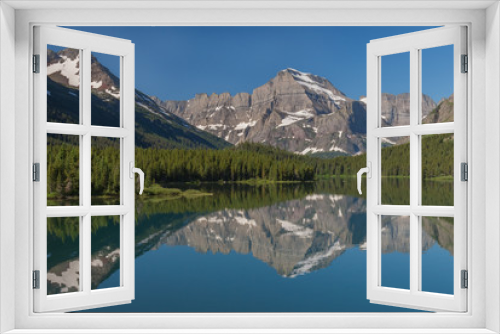 Fototapeta Naklejka Na Ścianę Okno 3D - Reflections of the rugged mountains towering above the forested shores can be seen in Swiftcurrent Lake on a beautiful summer day in Glacier National Park