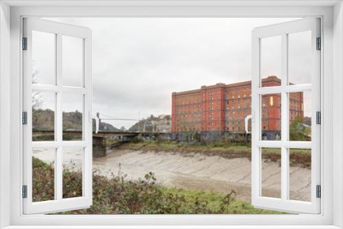 Fototapeta Naklejka Na Ścianę Okno 3D - The Avon river with mud after a storm, next to a red brick abandoned industrial building and a bridge in a cloudy winter in Bristol, United Kingdom