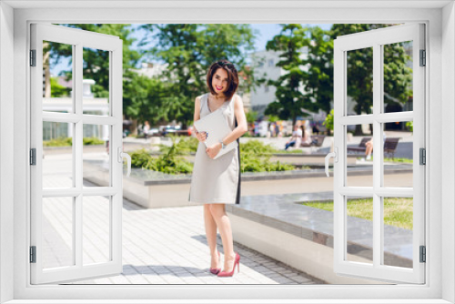 Pretty brunette girl in gray dress and vinous heels is standing in park in the city. She holds laptop and looks shy.