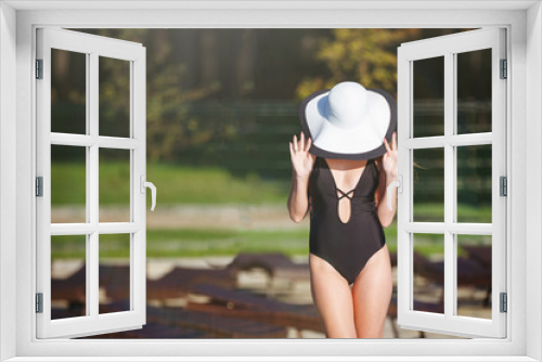 Fototapeta Naklejka Na Ścianę Okno 3D - Young woman in a black swimsuit and a beach hat near a pool with blue water in a resort hotel.