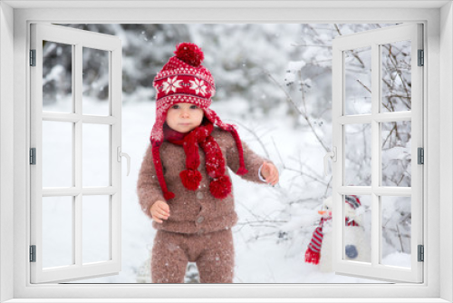 Portrait of a cute toddler baby dressed in a brown hand knitted jacket, pants, red hat and scarf, holding teddy and lantern, walks through the snowy park enjoying first snow blowing