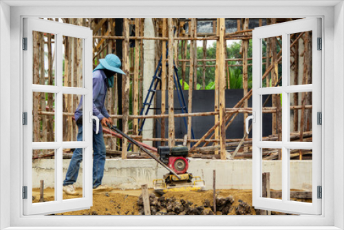 Fototapeta Naklejka Na Ścianę Okno 3D - worker at sand ground compaction with vibration plate compactor machine. with a background is the structure of the construction of the building. with cement base and eucalyptus wood