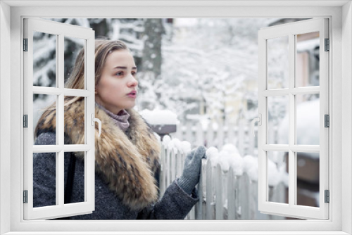 Stylishly dressed, beautiful girl in a fur coat, stands near the fence on the background of a residential building. The girl sadly looks at the windows of his house. Snowflakes fall on her shoulders.