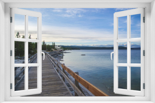 Fototapeta Naklejka Na Ścianę Okno 3D - Wooden pier on the beach of Yellowstone lake. Mountains view on background. Small forest on the side. Blue sky with some clouds and blue lake with geysers. 