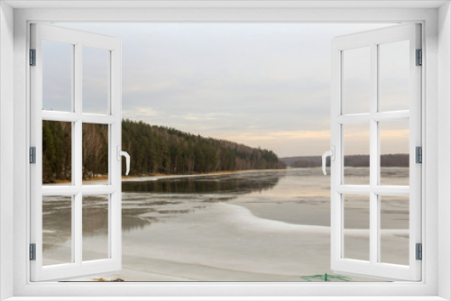 Fototapeta Naklejka Na Ścianę Okno 3D - View of the winter lake with an ice edge at the shore and low hills covered with forest