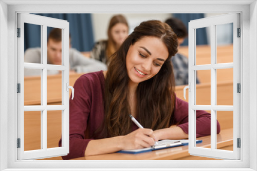 Pretty and beautiful brunette woman writing and smiling during lecture at university. Young female student learning and enjoying studying. Concept of education in modern collage.