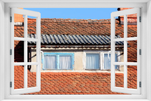Fototapeta Naklejka Na Ścianę Okno 3D - Roof windows with light blue and white dilapidated wooden frames surrounded with small roof tiles and rusted gutter next to two red brick chimneys on clear blue sky background