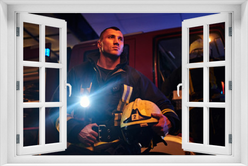 Fototapeta Naklejka Na Ścianę Okno 3D - The fire brigade came to the call at night. Handsome fireman wearing a protective uniform with a flashlight included, holding a helmet and looking sideways